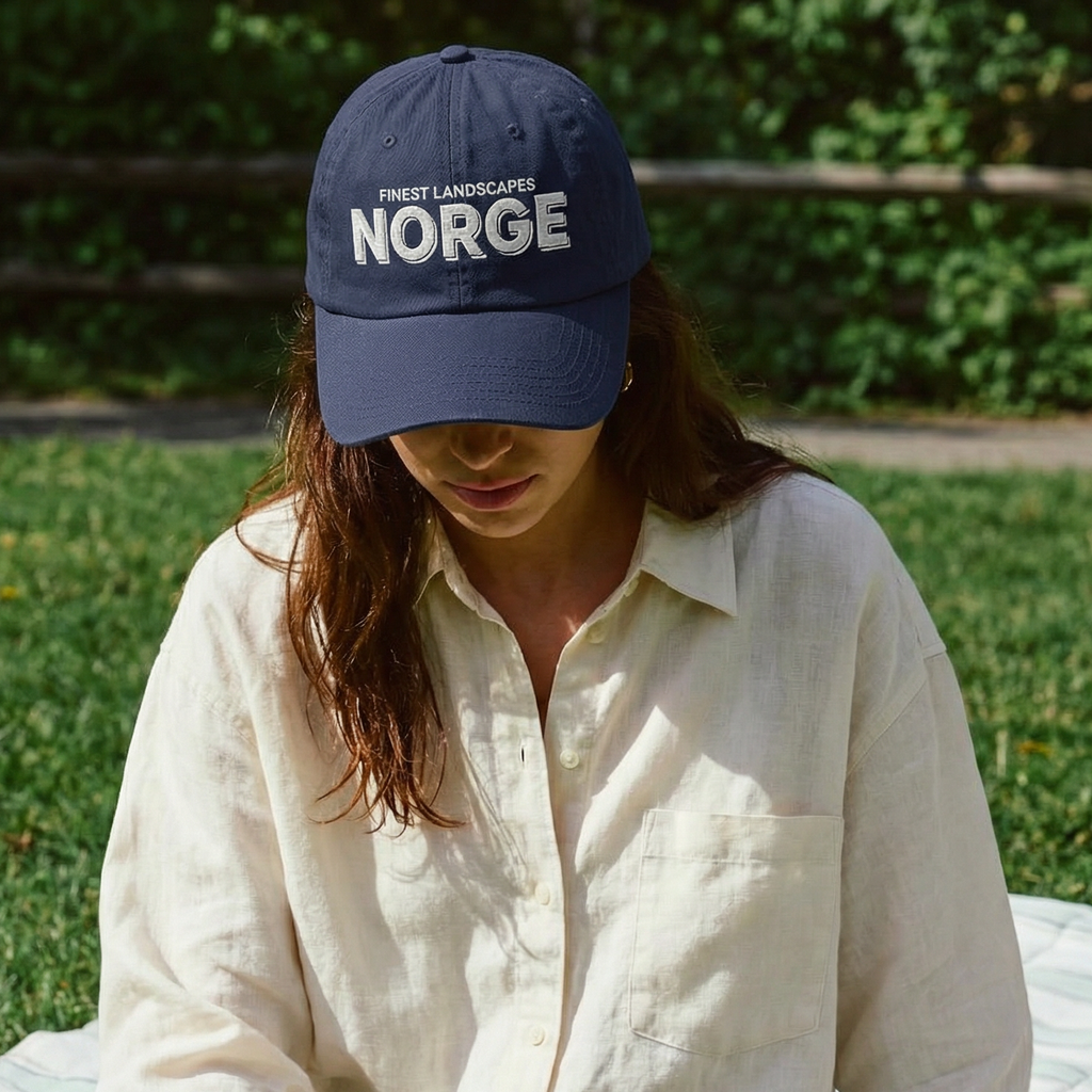 Person sitting on a blanket outdoors wearing a 'Norge' cap and holding a glass of wine.