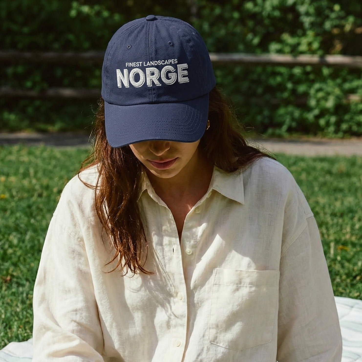 Person sitting on a blanket outdoors wearing a 'Norge' cap and holding a glass of wine.